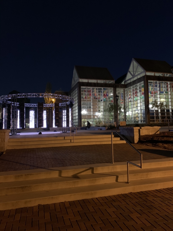 12 glowing LED panels are hung from a circular truss on an outdoor plaza next to a building comprised of glass panels that is glowing from the light within